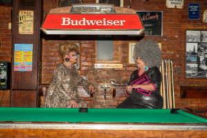 Drag performers Penny Tration and Molly Mormen sit and talk beside a pool table under a Budweiser sign at Rosie’s Tavern in Covington, Kentucky.
