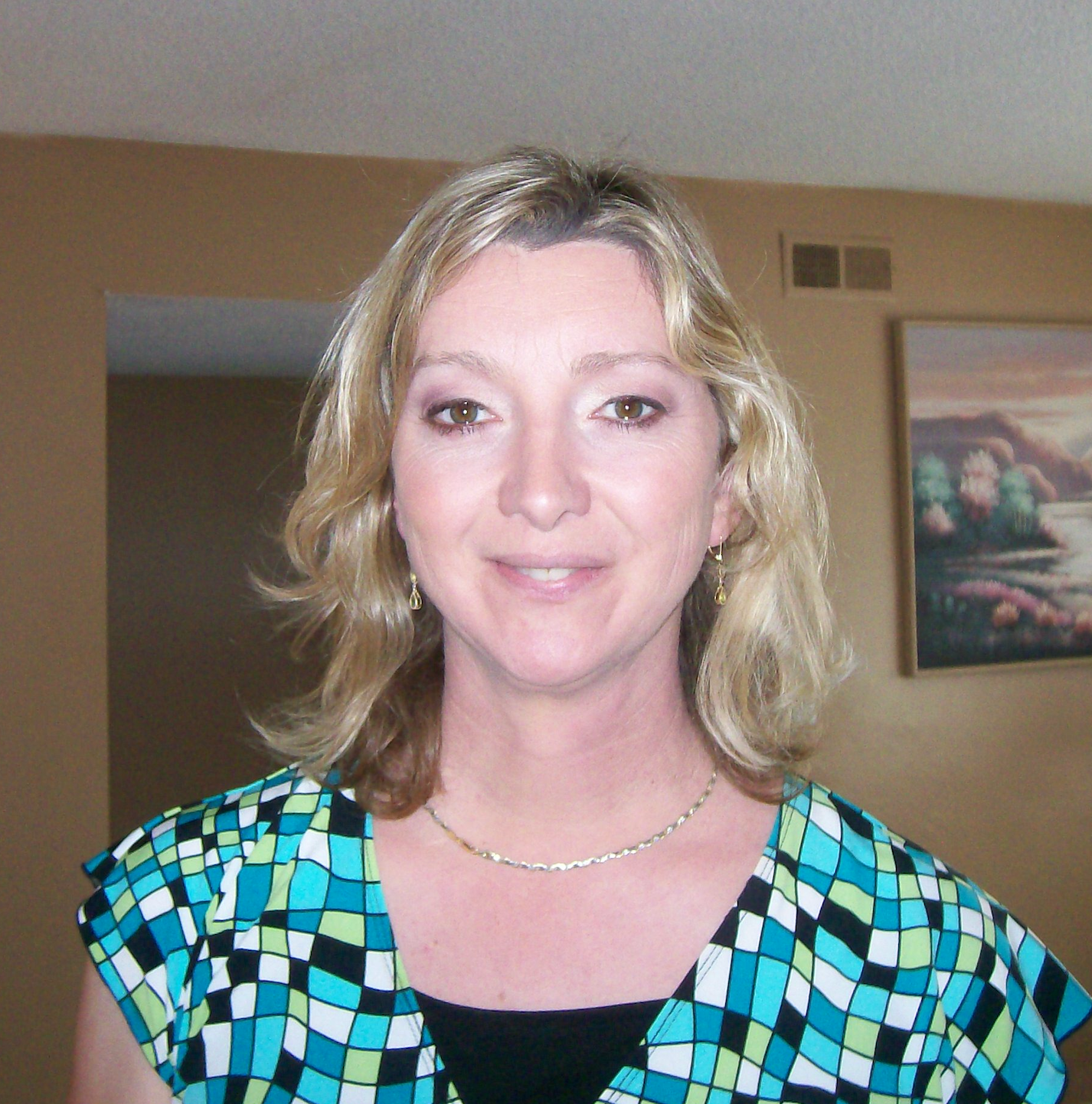 Heather Stevens smiling indoors, wearing a turquoise patterned blouse with a tan wall and painting behind her.