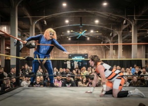 A performer in a blue bodysuit reaches toward an opponent in orange and white during a Power! Pride! Pro Wrestling match at Rhinegeist Brewery in Cincinnati, as the crowd cheers from ringside.