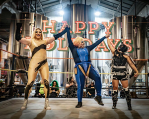 Three performers celebrate in the ring during a Power! Pride Pro Wrestling event at Rhinegeist Brewery in Cincinnati, raising their arms under a “Happy Holidays” sign as the crowd cheers behind them.
