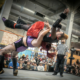 A drag wrestler in a red outfit is lifted into the air by another in purple and black during a match at a brewery, as a referee and crowd look on. Power! Pride! Pro Wrestling