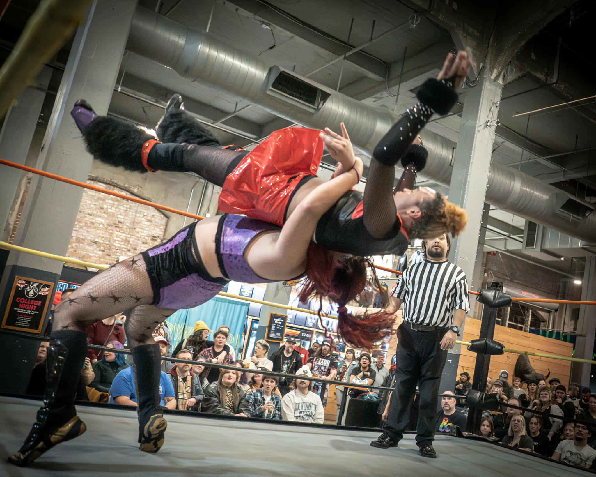 A drag wrestler in a red outfit is lifted into the air by another in purple and black during a match at a brewery, as a referee and crowd look on. Power! Pride! Pro Wrestling