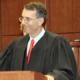 Judge Lewis Grant Paisley stands in a Kentucky courtroom wearing judicial robes and a red tie.