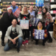 A group of people at Creative House of Art and Design in Covington, Kentucky, pose with their finished winter-themed paintings during a guided art class. The group stands in front of shelves filled with colorful art supplies and craft materials.