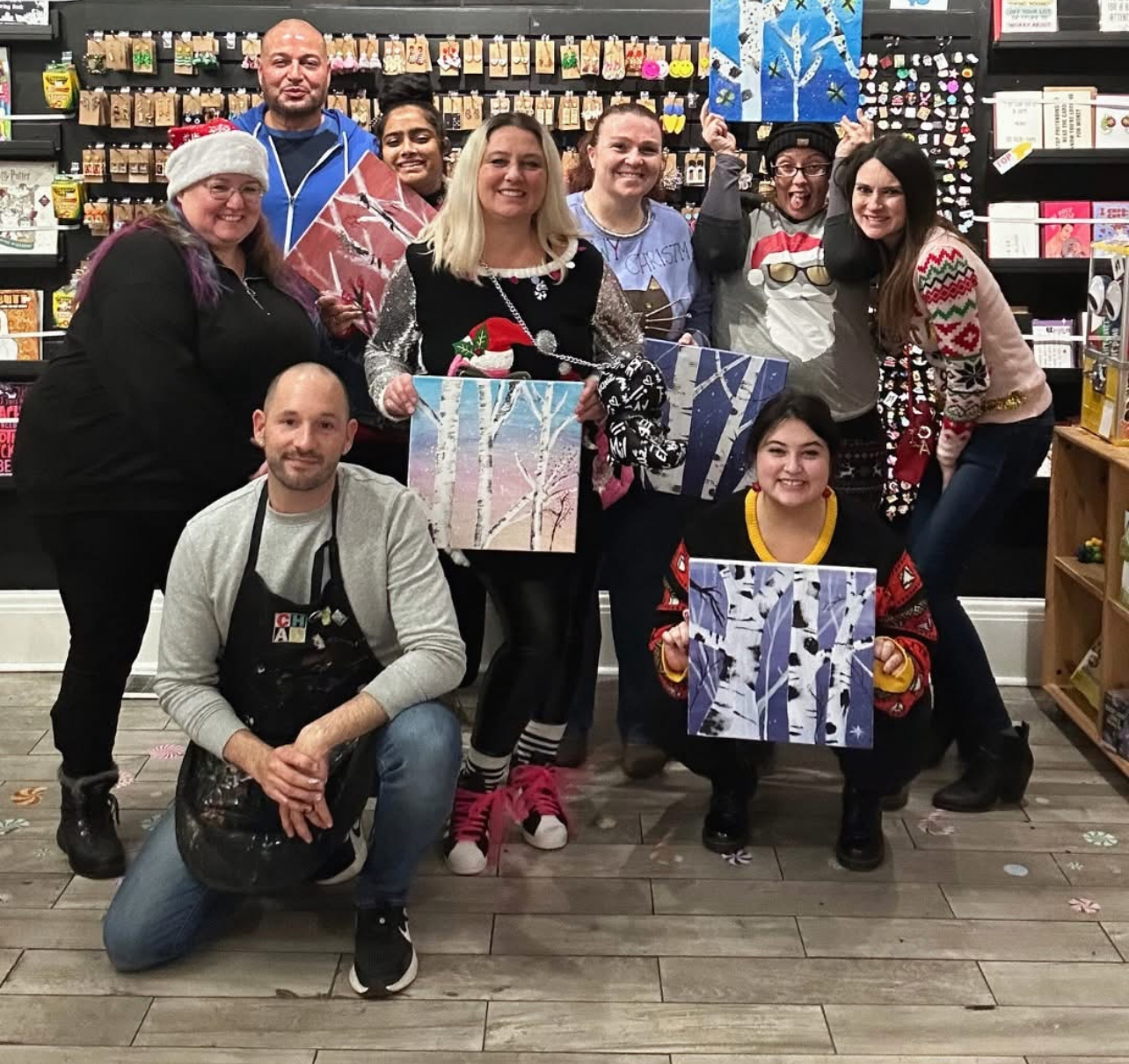 A group of people at Creative House of Art and Design in Covington, Kentucky, pose with their finished winter-themed paintings during a guided art class. The group stands in front of shelves filled with colorful art supplies and craft materials.