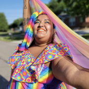 Sophia Harris Sophia Harris smiles brightly outdoors while holding a flowing rainbow-colored fabric above her head. She wears a vibrant multicolored outfit and stands in sunlight on a neighborhood street.