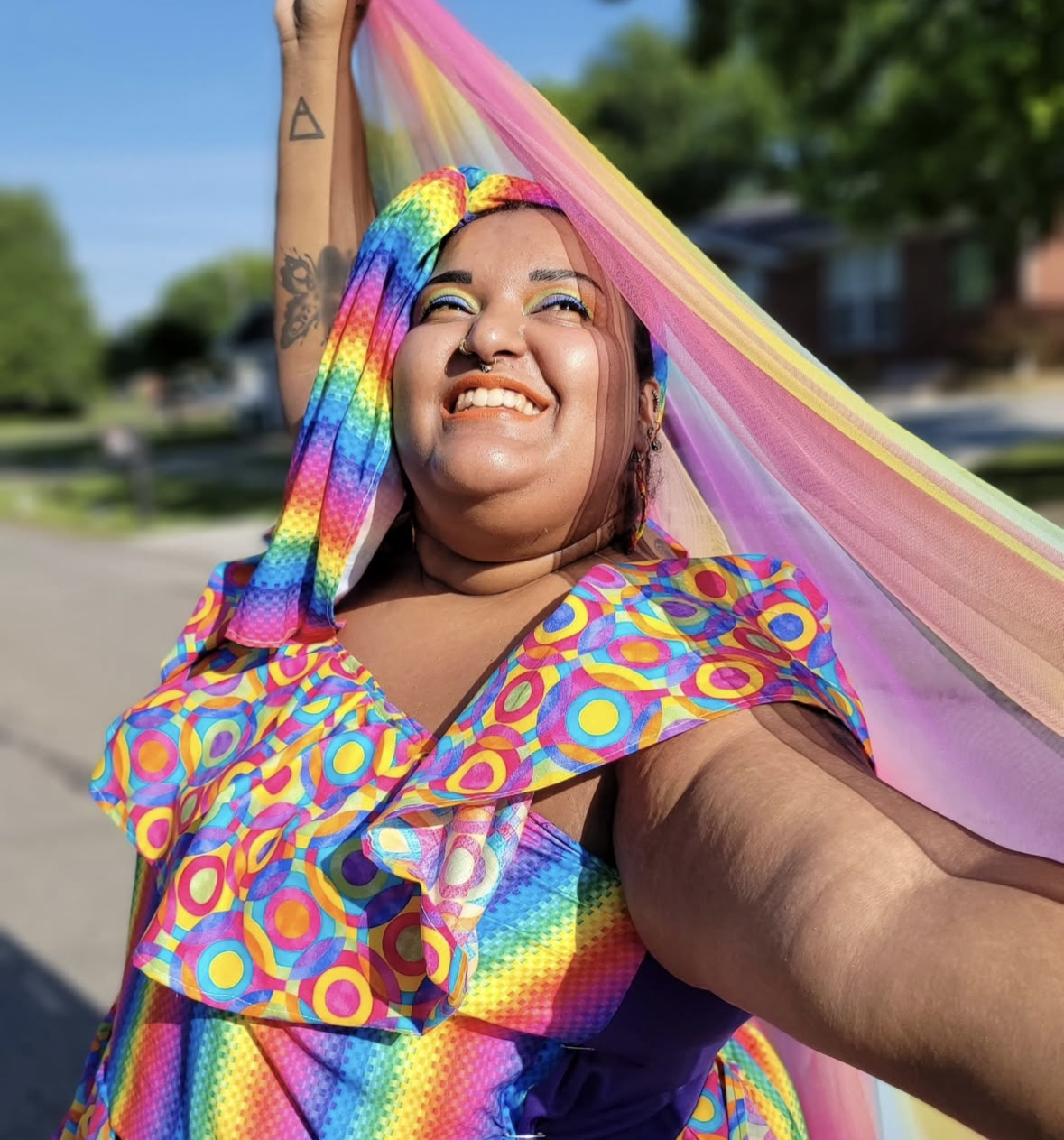 Sophia Harris smiles brightly outdoors while holding a flowing rainbow-colored fabric above her head. She wears a vibrant multicolored outfit and stands in sunlight on a neighborhood street.