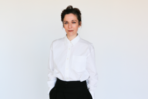 Jen Tullock stands against a plain white background wearing a white button-down shirt and black pants. The Kentucky-born actor and writer, known for Severance and her play Nothing Can Take You From the Hand of God, looks confidently at the camera with a relaxed posture.