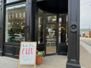 Exterior of Rekas Butchery and Delicatessen in Covington, Kentucky, with a sidewalk sign reading “We Are Open Today” in front of the shop’s black storefront and glass door.