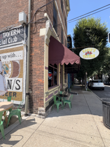Exterior of Rosie’s Tavern in Covington, Kentucky, showing its brick facade, vintage painted signage, maroon awning, and outdoor seating with green chairs.