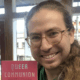 Davis Shoulders with long hair and glasses smiling indoors while holding up a pink book titled “Queer Communion: Religion in Appalachia.” Sunlight filters through the windows behind them, revealing part of a small-town street outside.