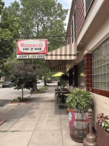Exterior of Jerry’s Jug House in Newport, Kentucky, featuring a vintage Wiedemann Beer sign, striped awning, and outdoor seating along the tree-lined sidewalk.