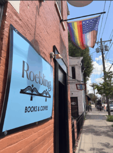Exterior of Roebling Books & Coffee in Newport, Kentucky, showing the shop’s blue sign on a brick wall and a Progress Pride flag with American stars hanging above the entrance on a sunny day.