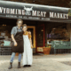 Wives Shelbi and Ashley Nation, known as the Nation wives, stand arm in arm in front of Wyoming Meat Market. They are smiling, wearing work aprons, and standing on the brick sidewalk beneath the shop’s black awning that reads “Wyoming Meat Market.” A bench and storefront windows are visible behind them.