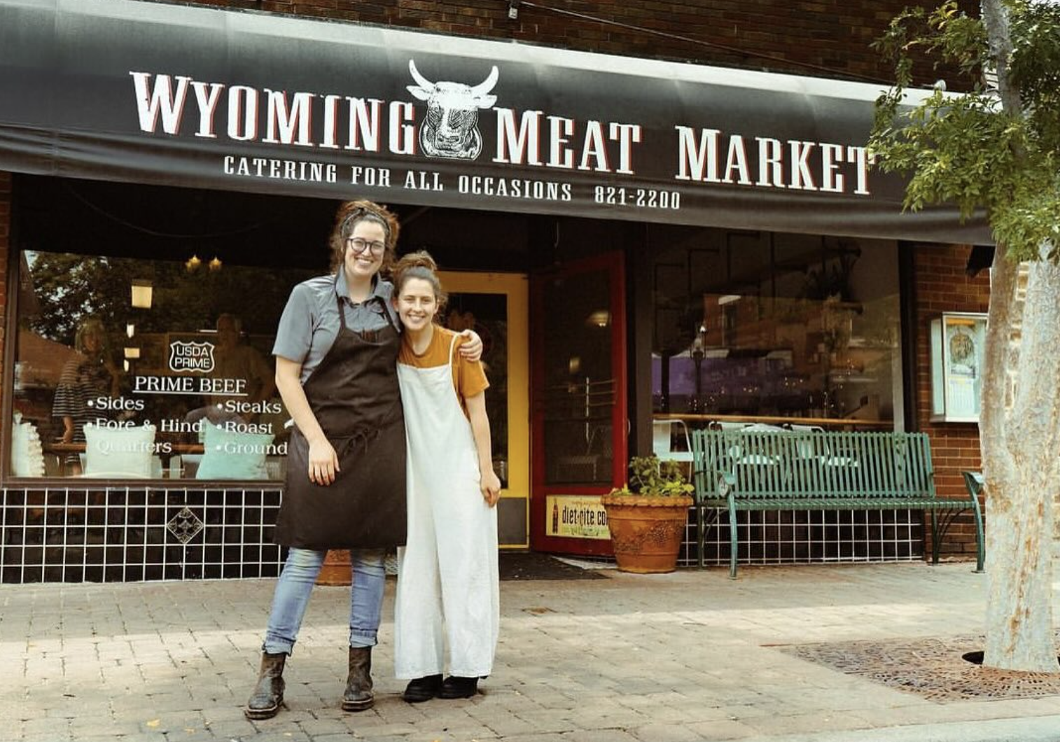 Wives Shelbi and Ashley Nation, known as the Nation wives, stand arm in arm in front of Wyoming Meat Market. They are smiling, wearing work aprons, and standing on the brick sidewalk beneath the shop’s black awning that reads “Wyoming Meat Market.” A bench and storefront windows are visible behind them.