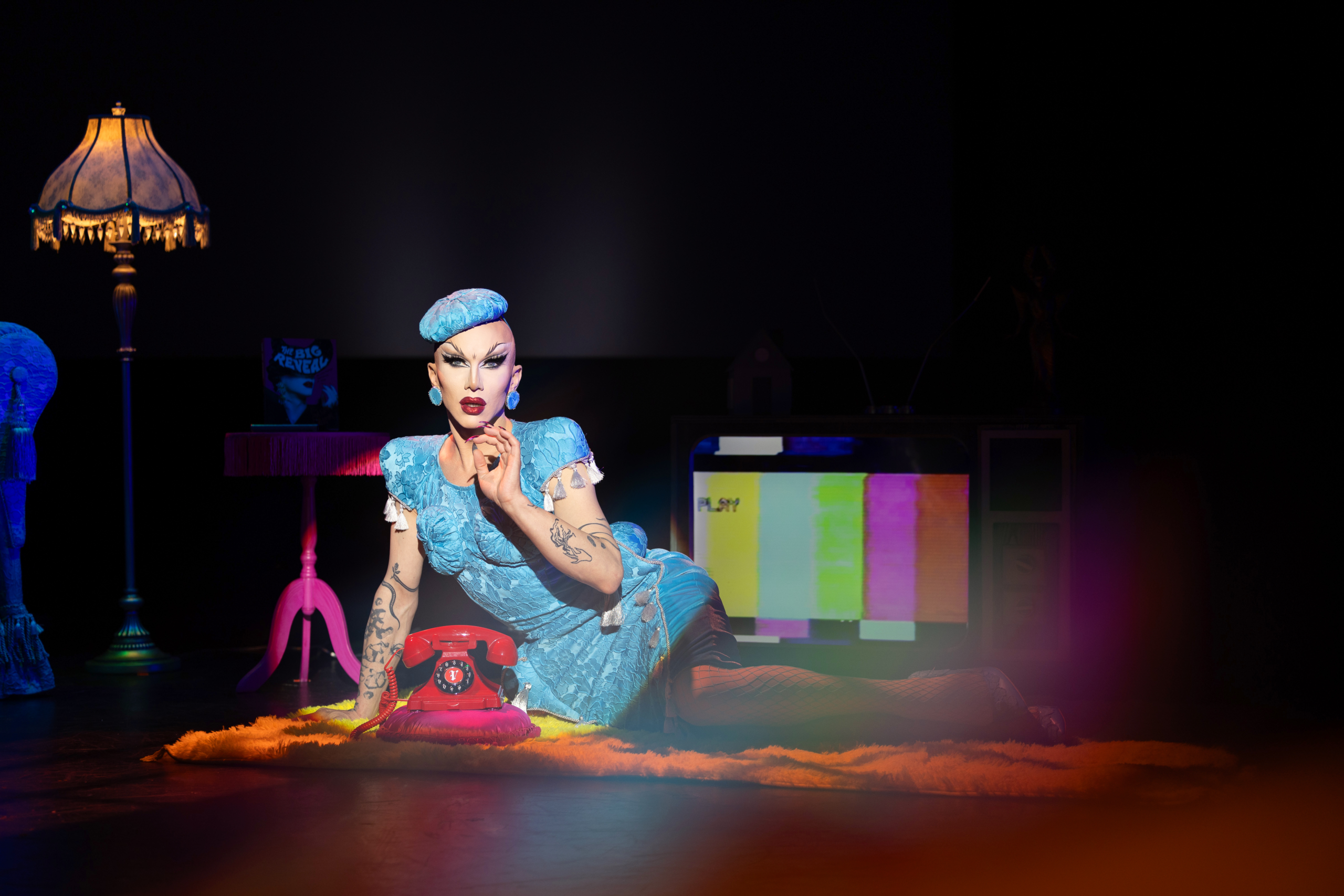 Sasha Velour poses in a blue outfit beside a red rotary phone during “The Big Reveal Live Show.”