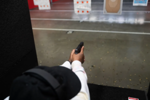 Xian Brooks wearing a black hat and white hoodie aims a handgun at paper targets inside an indoor shooting range.
