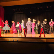 A lineup of drag performers stands across a brightly lit stage at the Slay Bells holiday show, wearing festive gowns, wigs, and glamorous costumes. Large red letters spell out “IVA” in front of them, with oversized Christmas ornaments decorating the background. Red curtains frame the stage as the performers pose and interact during the fundraiser for Moveable Feast Lexington.