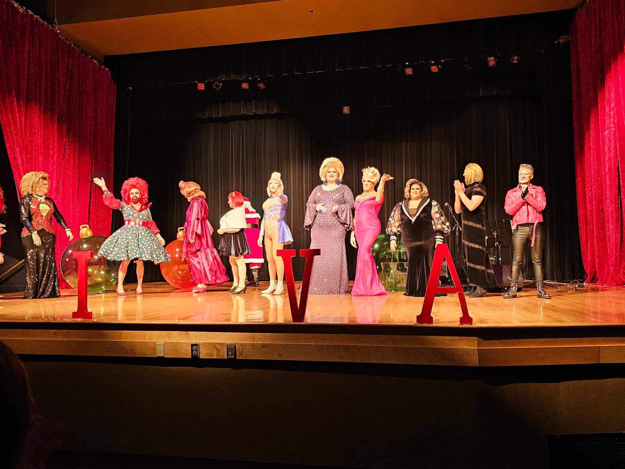A lineup of drag performers stands across a brightly lit stage at the Slay Bells holiday show, wearing festive gowns, wigs, and glamorous costumes. Large red letters spell out “IVA” in front of them, with oversized Christmas ornaments decorating the background. Red curtains frame the stage as the performers pose and interact during the fundraiser for Moveable Feast Lexington.