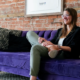 Emily Schubert, a licensed clinical social worker, sits on a purple couch inside Thrive Empowerment Center in Covington, Kentucky, a trauma-informed LGBTQ-affirming community space focused on safety, healing, and advocacy amid rising anti-LGBTQ+ violence in Northern Kentucky.