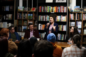 Policy expert Kara Stewart speaks to a packed audience at Queer Kentucky’s Queer Politics in the Bluegrass legislative preview panel in Newport, with panelists seated beside her inside Roebling Books.