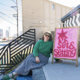 Kaleigh Basso sits outside Set & Setting, her queer independent bookstore in Louisville, beside a colorful hand-painted sign welcoming visitors to the shop.