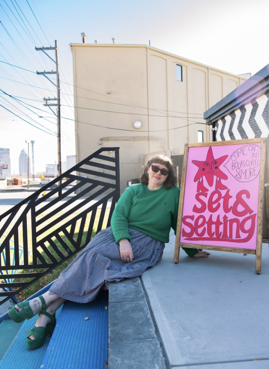 Kaleigh Basso sits outside Set & Setting, her queer independent bookstore in Louisville, beside a colorful hand-painted sign welcoming visitors to the shop.