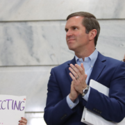 Gov. Andy Beshear, in a blue suit claps while standing inside a marble government building during a Fairness Rally, as people around him hold protest signs advocating for LGBTQ+ protections.