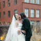 Heather Shaw and Lilly Jean Coiner embrace during their wedding portraits outdoors, with Coiner wearing a white wedding gown and veil and Shaw wearing a black suit while holding a bouquet of dark flowers. Brick buildings and tall grasses are visible in the background.