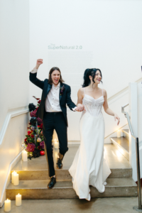 Heather Shaw and Lilly Jean Coiner walk hand in hand down a staircase during their wedding celebration. Shaw wears a black tuxedo and raises her fist in celebration, while Coiner wears a white wedding gown. Candles line the steps, with floral arrangements and gallery walls visible behind them.