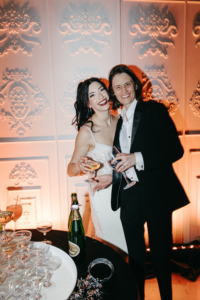 Heather Shaw and Lilly Jean Coiner pose together at their wedding reception, smiling while holding champagne glasses. Shaw wears a white wedding dress, and Coiner wears a black tuxedo, standing in front of an ornate white wall with warm lighting and a table of stacked coupe glasses nearby.