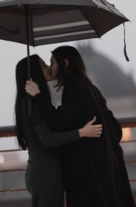 Heather Shaw and Lilly Jean Coiner share a kiss under a black umbrella on a rainy day, holding each other closely. Both are dressed in dark clothing, standing outdoors with a soft, overcast sky behind them, creating an intimate and romantic moment.