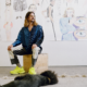 John Brooks seated on a wooden box in a gallery, wearing a blue shirt and bright green shoes, with large figurative drawings displayed on the wall behind them and a black dog lying on a rug in the foreground.