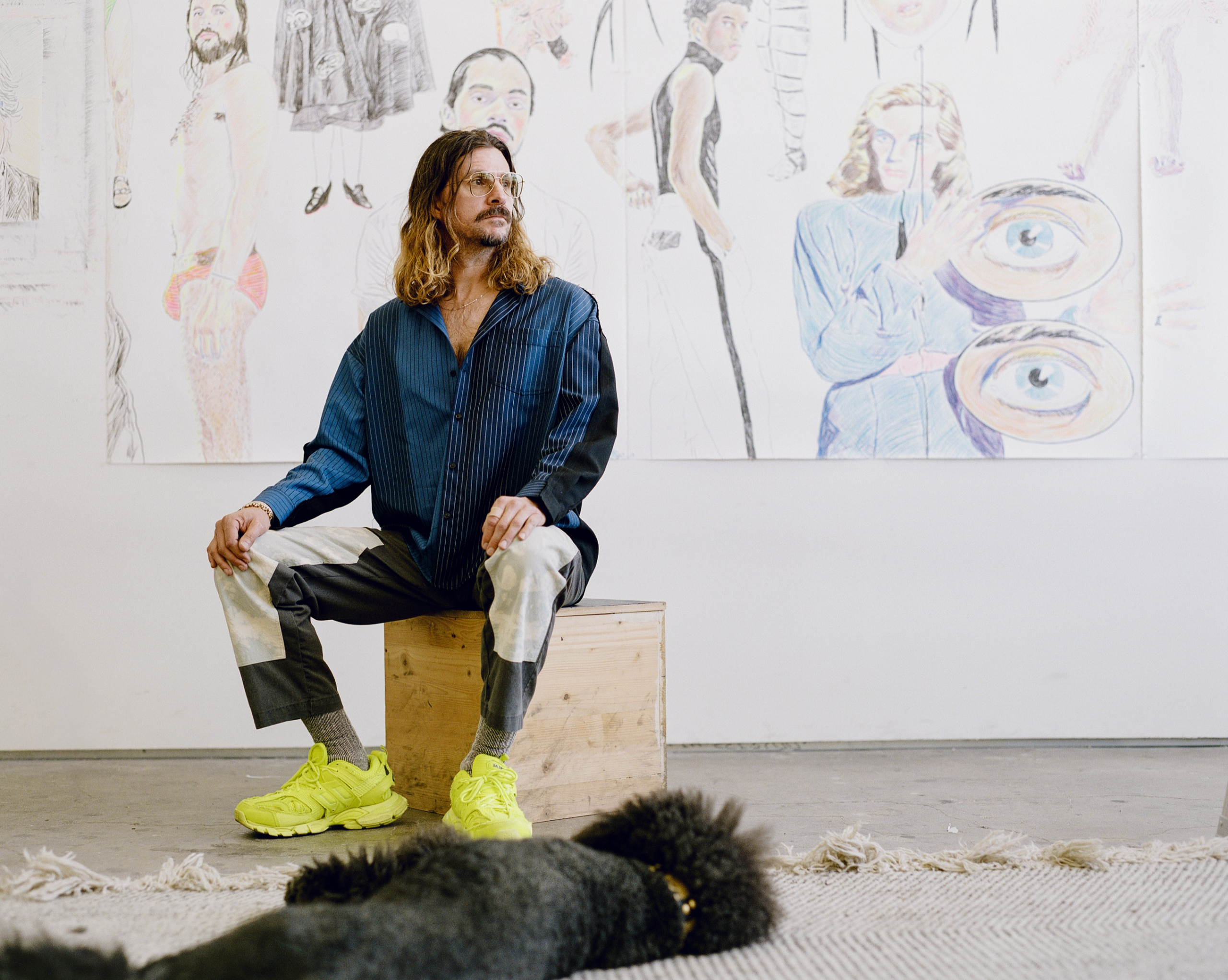 John Brooks seated on a wooden box in a gallery, wearing a blue shirt and bright green shoes, with large figurative drawings displayed on the wall behind them and a black dog lying on a rug in the foreground.