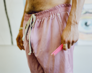 Close-up of artist John Brooks’ torso and hands, wearing pink drawstring pants and holding two pink pencils, with a blurred artwork visible in the background.
