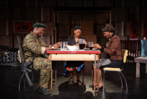 Three actors sit around a small table onstage during a theater performance. One wears a military uniform with a rifle resting beside his chair, while the two others wear civilian clothing. They share a meal together under warm stage lighting, with a modest set and framed artwork in the background.