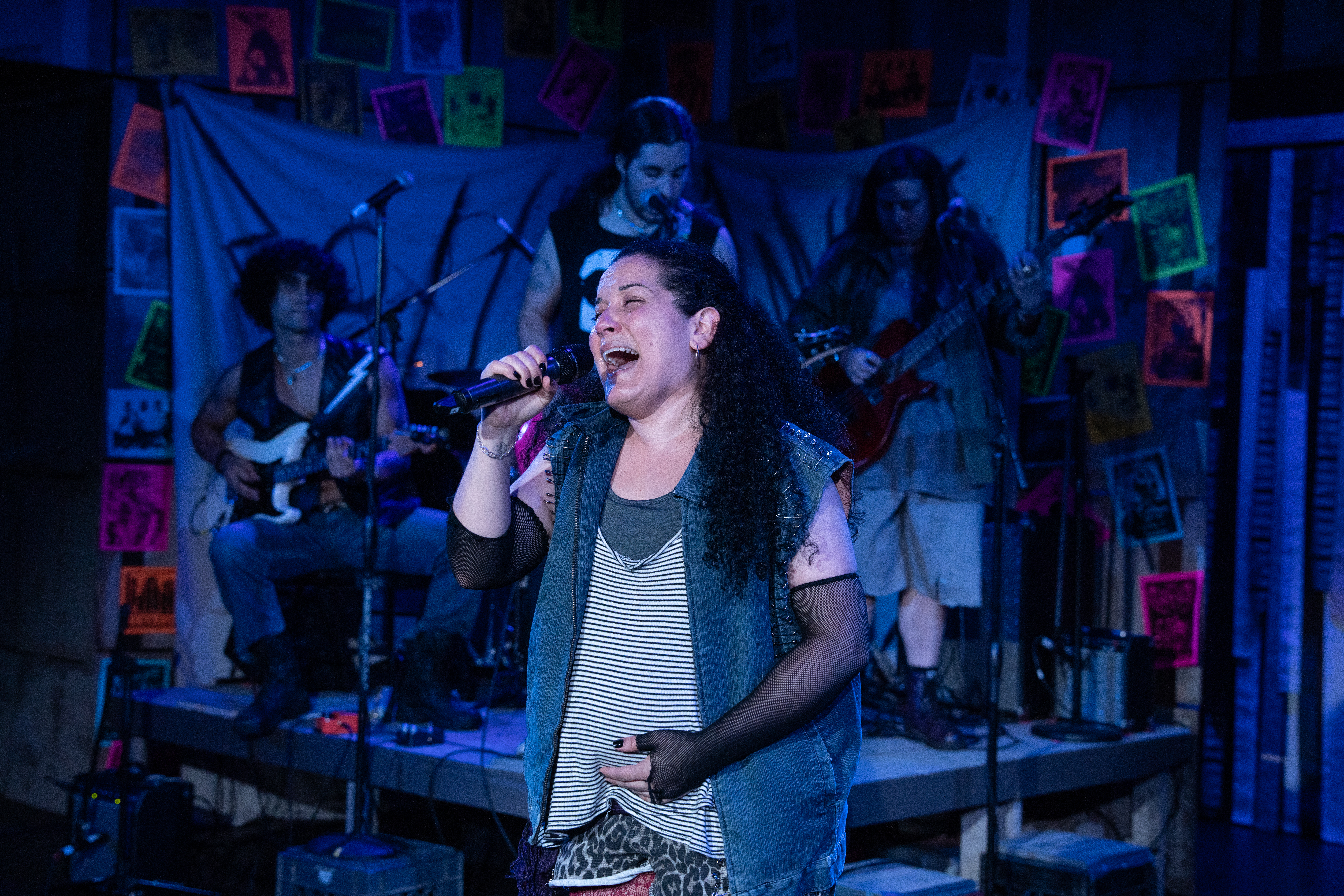 A performer sings passionately into a handheld microphone on a small stage under blue lighting, backed by a live band with guitars and drums. Colorful posters hang behind the musicians, creating a vibrant, community theater atmosphere.