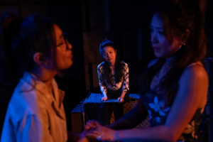 Three actors perform an intimate scene onstage under dramatic blue and red lighting. Two stand close together in the foreground holding hands, while a third sits in the background watching, creating a tense and emotional moment within a minimalist theater set.