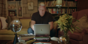 Author David Dominé sits in a warmly lit living room in Old Louisville, working on a laptop at a coffee table stacked with books, with disco balls and a bookshelf in the background.