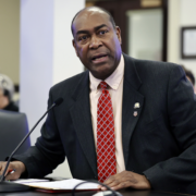 Sen. Donald Douglas, R-Nicholasville, speaks at a microphone during a Kentucky Senate Committee on Health Services meeting in Frankfort, presenting Senate Bill 72 (SB 72). He sits at a desk with papers in front of him, wearing a dark suit and red patterned tie, as attendees listen in the background.