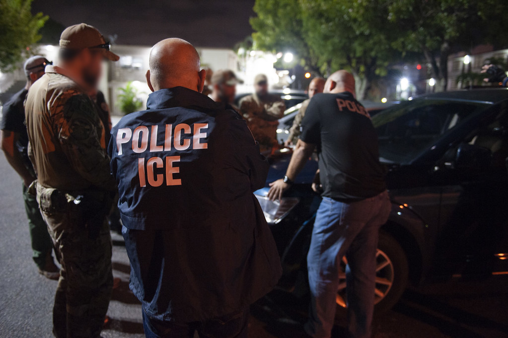 U.S. Immigration and Customs Enforcement officers and law enforcement agents gather around a vehicle during an immigration enforcement operation at night.