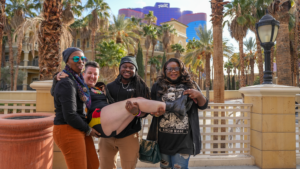 Four LGBTQ conference attendees laugh and pose outside the Rio hotel in Las Vegas, one person being lifted by the group, with palm trees in the background.
