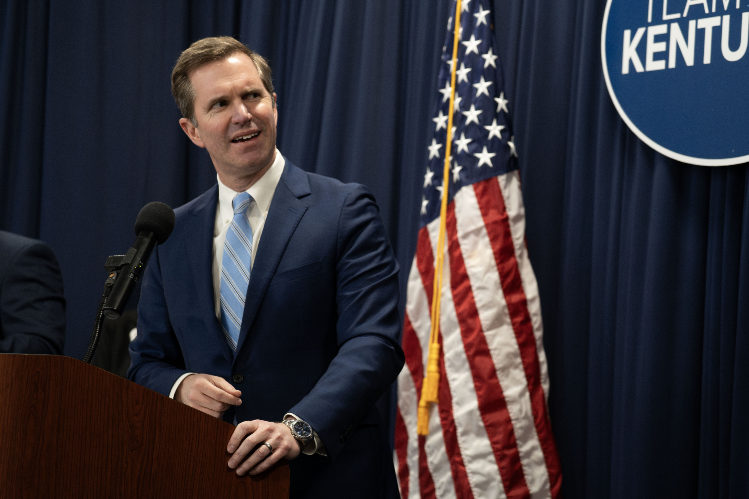 Kentucky Gov. Andy Beshear speaks at a podium during a press conference, with an American flag and Team Kentucky branding visible in the background.