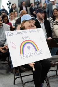 A smiling woman in a light blue cap holds a handmade sign reading "Make America Great Again With LGBTQ+ Pride" featuring a rainbow and the words "Love Wins" and "Love is Love" during a crowded public hearing.